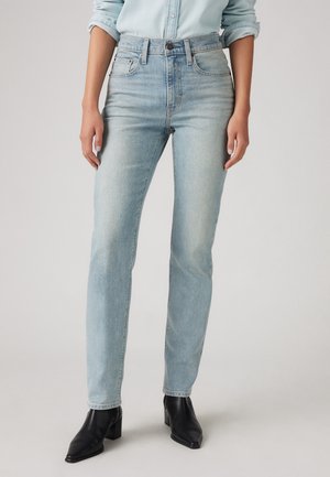 Femme portant un jean slim taille haute bleu clair, une chemise bleu clair boutonnée et des bottines noires à talons, debout devant un fond uni.