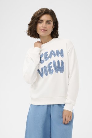 Kobieta o krótkich brązowych włosach, ubrana w biały sweatshirt z napisem „OCEAN VIEW” niebieskimi literami i jasno-niebieskie szerokie spodnie.