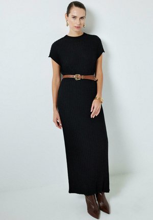 Vestido negro de punto acanalado con mangas cortas, cintura ajustada acentuada por un cinturón marrón, combinado con botas de tacón marrón con punta.