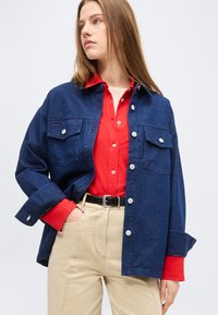 Noor naine, kellel on pikad juuksed, seljas punane nööpidega särk ja tumesinine teksajakk, beežid püksid ning must vöö, vaatab vasakule.