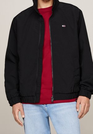 Schwarze Bomberjacke mit Stehkragen, Reißverschluss vorne, elastischen Bündchen und Saum; getragen über einem roten Shirt, kombiniert mit hellblauen Jeans.