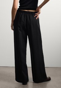 Pantaloni neri a gamba larga con vita arricciata, realizzati in un tessuto liscio e testurizzato, con un fit rilassato, abbinati a scarpe scure con punta chiusa.