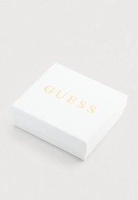 Λευκό κουτί δώρου με χρυσό λογότυπο "GUESS", τετράγωνο σχήμα, λεία υφή, κλειστό με καμία ορατή είσοδο ή επιπλέον σχεδιαστικά στοιχεία.