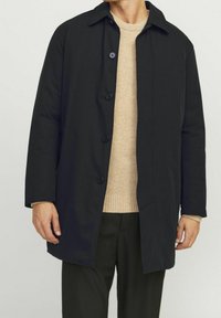 Černý kabát na knoflíky s plochým límcem, dlouhými rukávy a rovný řez. Nosený přes světle béžový svetr a tmavé kalhoty.