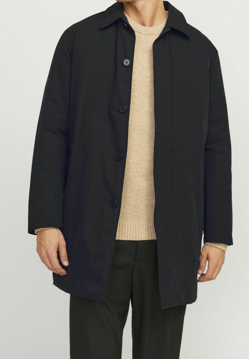 Černý kabát na knoflíky s plochým límcem, dlouhými rukávy a rovný řez. Nosený přes světle béžový svetr a tmavé kalhoty.