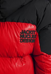 Rød og sort pufferjakke med quiltet tekstur; har sort broderet tekst "JACKIT NUCL1AR DESIGN" på den røde sektion.