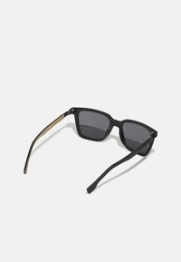 Svarte solbriller med firkantet ramme. Matt finish med grå linser. Stengene har en sandfarget aksent. Elegante og moderne design.