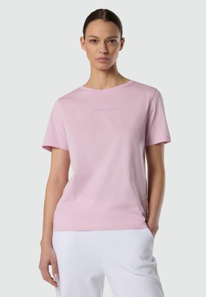 Donna con capelli scuri legati indietro che indossa una maglietta rosa chiaro a maniche corte e pantaloni bianchi, in piedi con una mano in tasca.