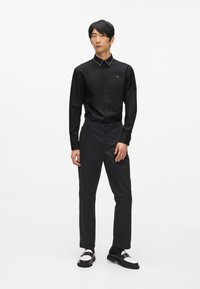 Fekete, gombos ing texturált gallérral, fehér szegecsekkel díszítve, fekete nadrággal és fekete-fehér cipőkkel párosítva, egy sima háttér előtt áll.