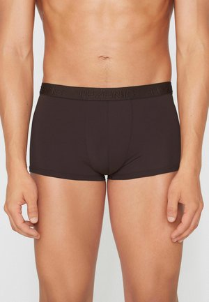 Männlicher Oberkörper, der schwarze, taillierte Boxershorts mit einem gebrandeten Bund trägt, steht vor einem schlichten hellen Hintergrund.