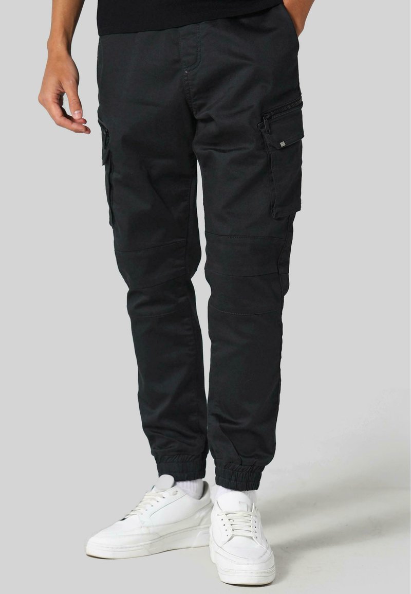 Pantalones cargo negros con múltiples bolsillos, puños elásticos y una textura suave y mate, combinados con zapatillas blancas.