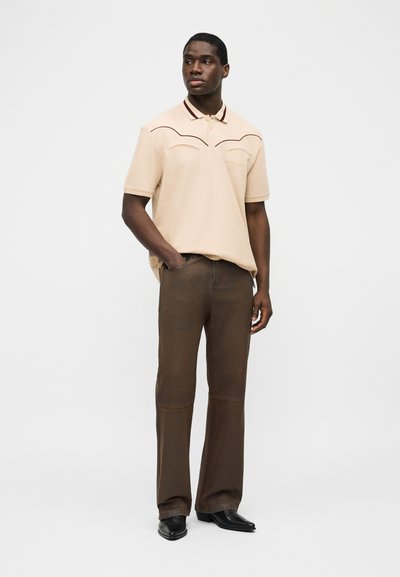 Homme portant un polo beige à manches courtes avec des bordures marron foncé, un pantalon droit marron, des chaussures noires pointues, debout avec une main dans la poche.