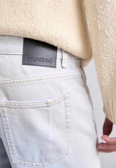 White denim jeans with a black leather tag labeled "Hundred." The fabric has a smooth texture and features standard back pockets.