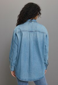 Lichtblauwe denimshirt met een losse pasvorm, kraag, lange mouwen en een rugplooidetail, voorzien van zichtbare stiksels en een afgeronde zoom.