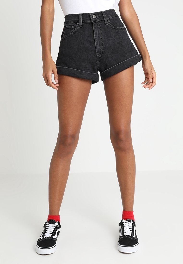 Femme portant un short en denim noir taille haute avec des ourlets retroussés, des chaussettes rouges aux chevilles et des baskets noires et blanches, debout devant un fond blanc.