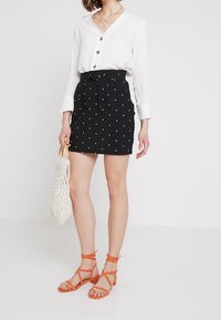Falda negra con lunares, con una cintura ceñida y un cinturón, combinada con una camisa blanca de botones. Sandalias de color tostado y un bolso de mimbre completan el look.