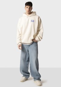 Felpa color crema con tasca frontale e una toppa grafica. Abbinata a jeans baggy azzurri e sneaker bianche.