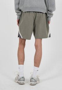 Personne portant un short gris avec des panneaux latéraux blancs et gris foncé, des chaussettes blanches montantes, des baskets grises et un pull gris, debout sur un fond blanc.