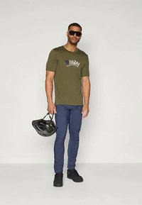 Camiseta de manga curta verde-azeitona com logotipo, calças justas azuis-marinho e ténis pretos. Segurando um capacete preto. Usando óculos de sol.