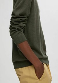 Olivgrön tröja med rundad hals och ribbade ärmslut. Mjuk stickad textur, något avslappnad passform, tillsammans med ljusbeiga byxor.