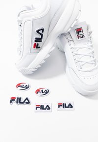 Sneakers bianchi FILA con logo ricamato, tomaia testurizzata e suole in gomma. Include cinque toppe con vari design di branding FILA.