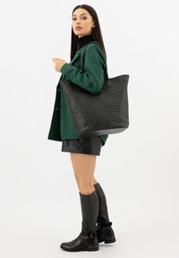 Borsa tote nera intrecciata con superficie strutturata e forma solida, dotata di manici lunghi e fondo liscio. Indossata con un cappotto verde sopra un completo nero.