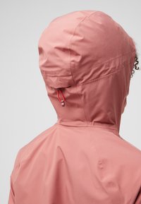 Giacca impermeabile rosa chiaro con cappuccio aderente, cordino elastico regolabile e texture liscia; particolare cucitura visibile sul retro.