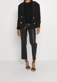 Blazer noir avec boutons dorés, pull noir orné en dessous, pantalon en simili cuir, et talons noirs à brides. Textures lisses et coupe ajustée.