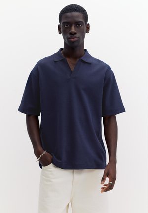 Junger Mann steht, trägt ein marineblaues Poloshirt mit kurzen Ärmeln, weiße Hose, silbernes Armband und blauen Steinring, vor einem einfarbigen weißen Hintergrund.