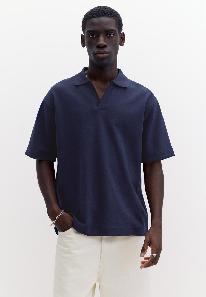 Jeune homme debout, portant un polo bleu marine à manches courtes, un pantalon blanc, un bracelet en argent et une bague avec une pierre bleue, sur un fond blanc uni.