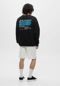 Czarna bluza z kapturem z niebieską grafiką cegieł na plecach, z białym napisem. Materiał: mieszanka bawełny. Noszona z białymi szortami i czarnymi butami.