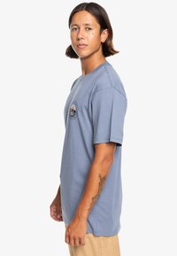 Light blue cotton T-shirt with short sleeves and a round neck. Features a circular graphic emblem on the left chest area.