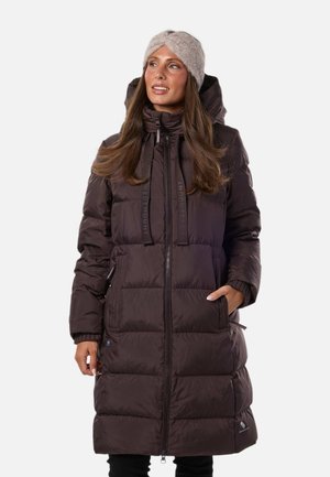 Mujer con un abrigo de invierno largo acolchado de color marrón oscuro con capucha y una banda para la cabeza beige de punto, de pie con una mano en el bolsillo.