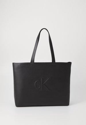 Černá kožená kabelka přes rameno se dvěma uchy, s vyraženým logem "cK" na přední straně. Hladká textura a minimalistický design.