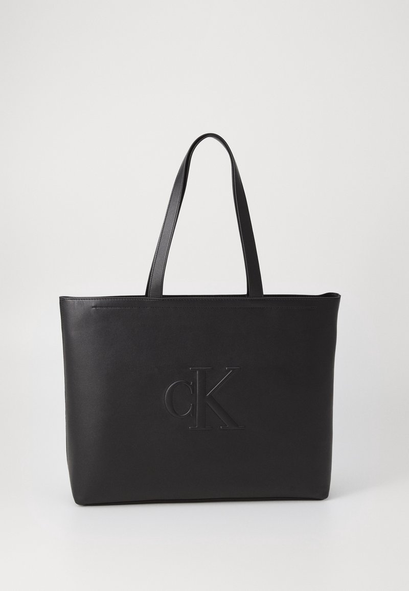 Černá kožená kabelka přes rameno se dvěma uchy, s vyraženým logem "cK" na přední straně. Hladká textura a minimalistický design.