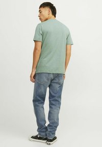 Camiseta verde de manga corta, tela ligera, cuello redondo. Combinada con jeans azul desgastados y zapatillas negras con acentos blancos.