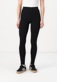 Zwarte legging met een gladde, rekbare stof, hoge tailleontwerp en enkel lengte, gecombineerd met zwart-witte sneakers.