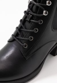 Botín de cuero negro que presenta puntera redonda, superficie texturizada, ojetes de metal y cordones redondos; tiene un tacón bajo y grueso para mayor estabilidad.