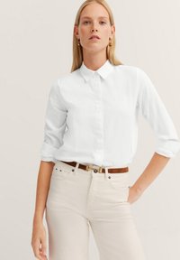 Weißes Hemd mit langen, hochgerollten Ärmeln aus strukturiertem Stoff, kombiniert mit hellen Jeans und einem braunen Gürtel.