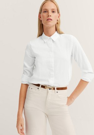 Weißes Hemd mit langen, hochgerollten Ärmeln aus strukturiertem Stoff, kombiniert mit hellen Jeans und einem braunen Gürtel.