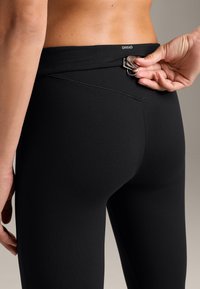 Leggings negre dintr-un material neted și elastic. Prezintă un buzunar discret pe spate cu un clapetă metalică și un logo la talie.
