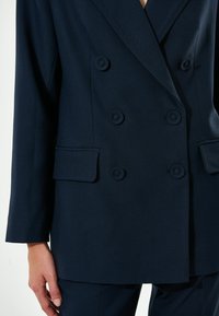 Blazer doppiopetto blu navy in tessuto testurizzato, con dettagli in bottone e due tasche frontali. Vestibilità su misura con maniche lunghe e rever a punta.