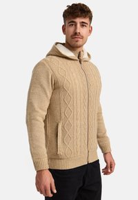 Sudadera de punto beige con patrón de trenzas texturizado, cierre de cremallera y bolsillos laterales. El interior tiene un suave forro contrastante en crema.
