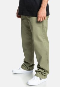 Olivengrønne oversized bukser, bomuldsstof, lige ben-design, manchetter ved anklerne, parret med hvide sneakers.