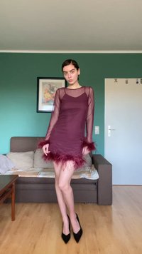 Vinröd klänning med långa, genomskinliga ärmar och fjäderdetaljer vid fållen och ärmsluten. Figursydd silhuett med slät textur och lager på lager-design.