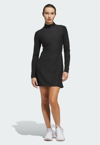 Zwarte langmouw jurk met een ritskraag, slim fit en zachte textuur, gecombineerd met witte sportschoenen.