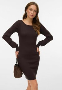 Haljina od rebrastog pletiva tamno smeđe boje, uskog kroja s dugim rukavima, dekoltea sa zaobljenim izrezom i bočnim prorezom, uparena s smeđom torbom od brušene kože.