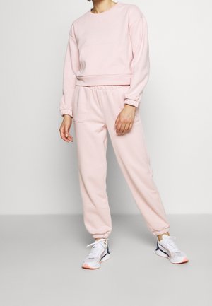 Person trägt ein hellrosa Sweatshirt und passende Jogginghosen sowie weiße und marineblaue Turnschuhe und steht vor einem schlichten hellen Hintergrund.