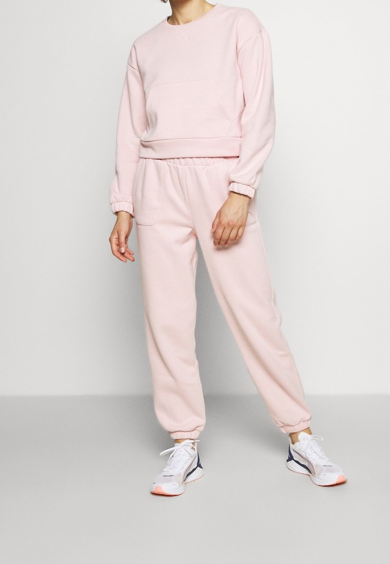 Personne portant un sweat-shirt rose clair et un pantalon de jogging assorti avec des baskets blanches et bleu marine, debout devant un fond clair et uni.