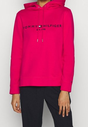 Osoba nosząca jasnoróżową bluzę z kapturem Tommy Hilfiger z logo oraz czarne spodnie, stojąca na jednolitym jasnym tle.
